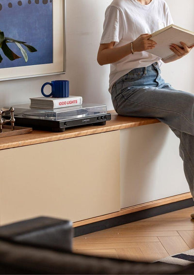Person sitting on a cabinet reading a book with a record player and decor items in the background.