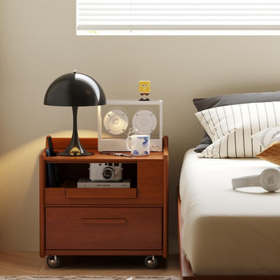 Wooden nightstand with lamp, speakers, and mug next to a bed with pillows and a window in the background.
