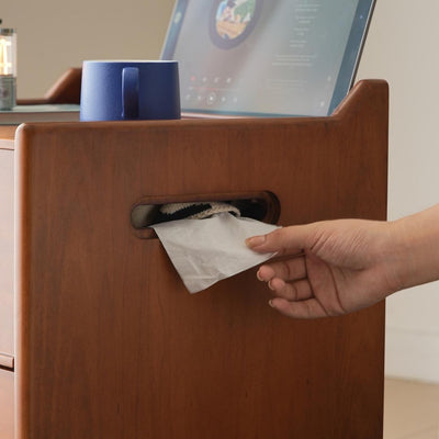 Hand pulling a paper towel from a wooden holder with a laptop in the background